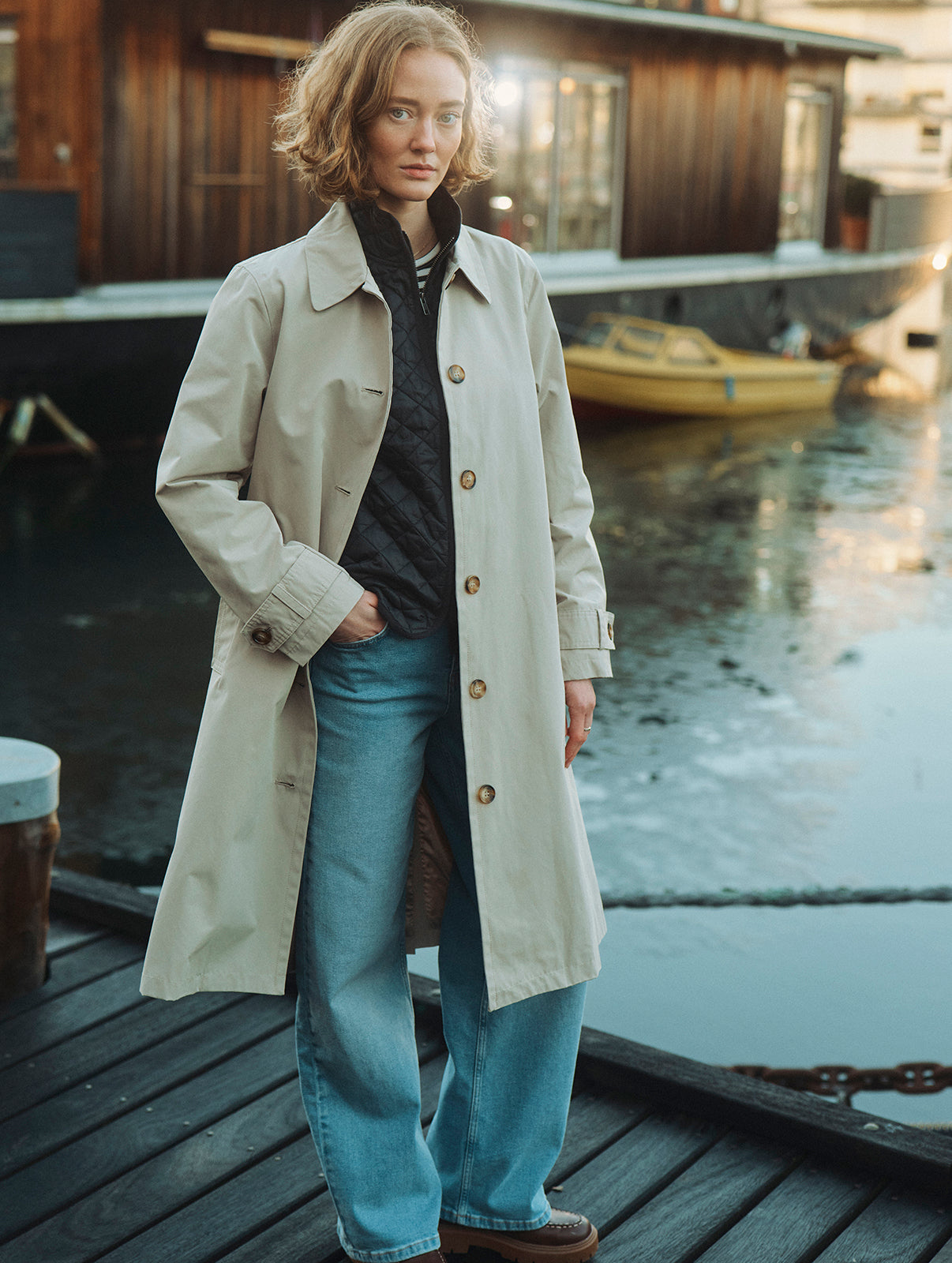 Kvinde i beige trenchcoat og lyse jeans står på en træbro ved vandet foran en husbåd.