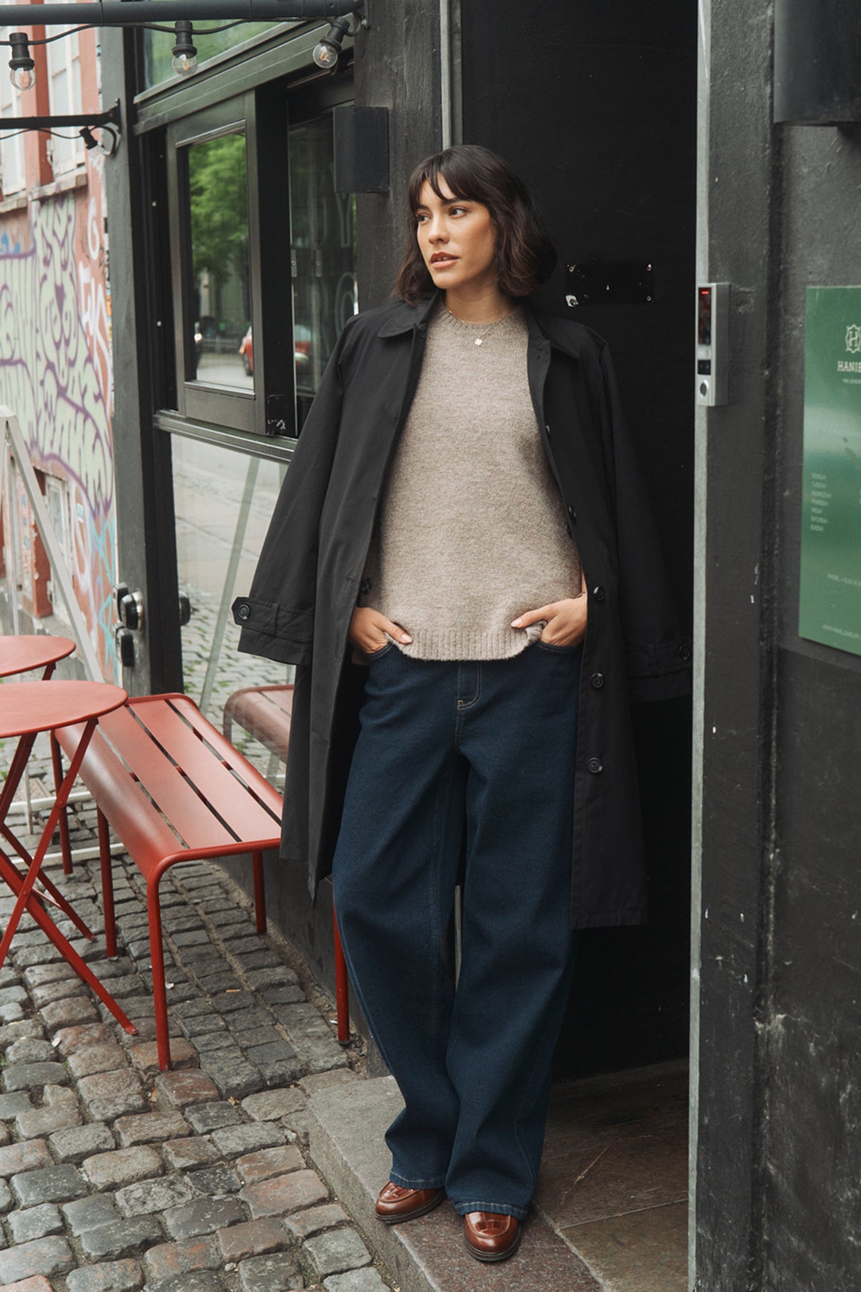 Oversize sort trenchcoat med lommer over beige strik og mørkeblå løstsiddende bukser.