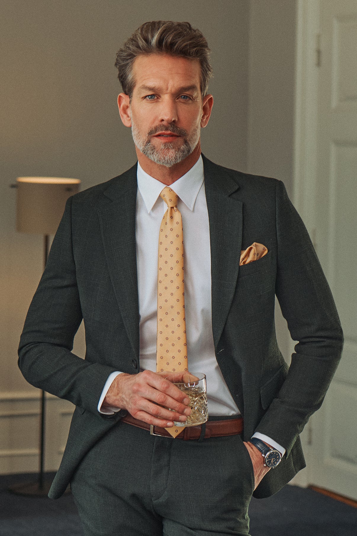 Mørkegrå tidløs uldblandingblazer med klassisk snit og sidelommer, stylet med hvid skjorte og lysegul slips.