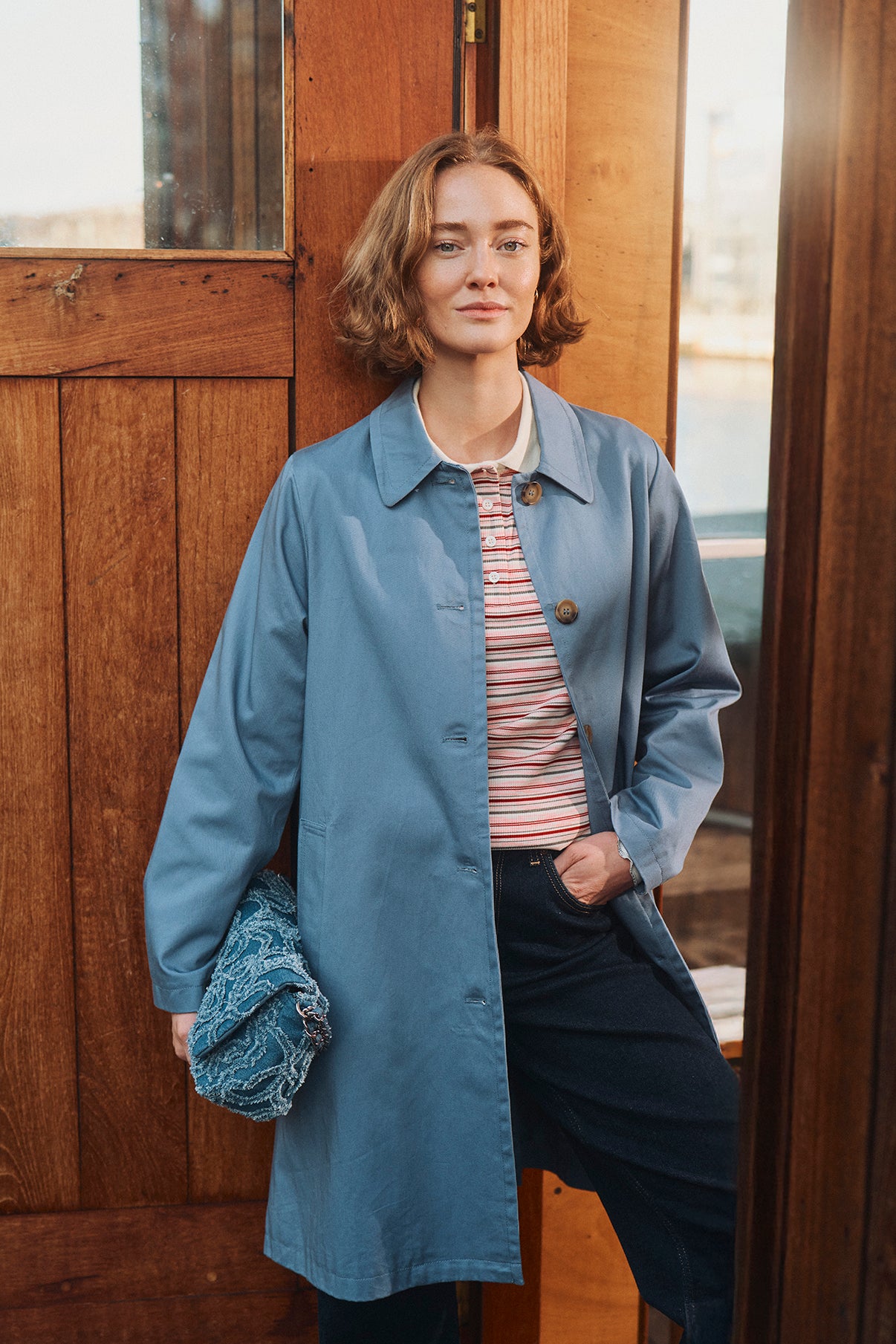 Lang, blå trenchcoat med knaplukning og sidelommer, styling med stribet top og mørke bukser.