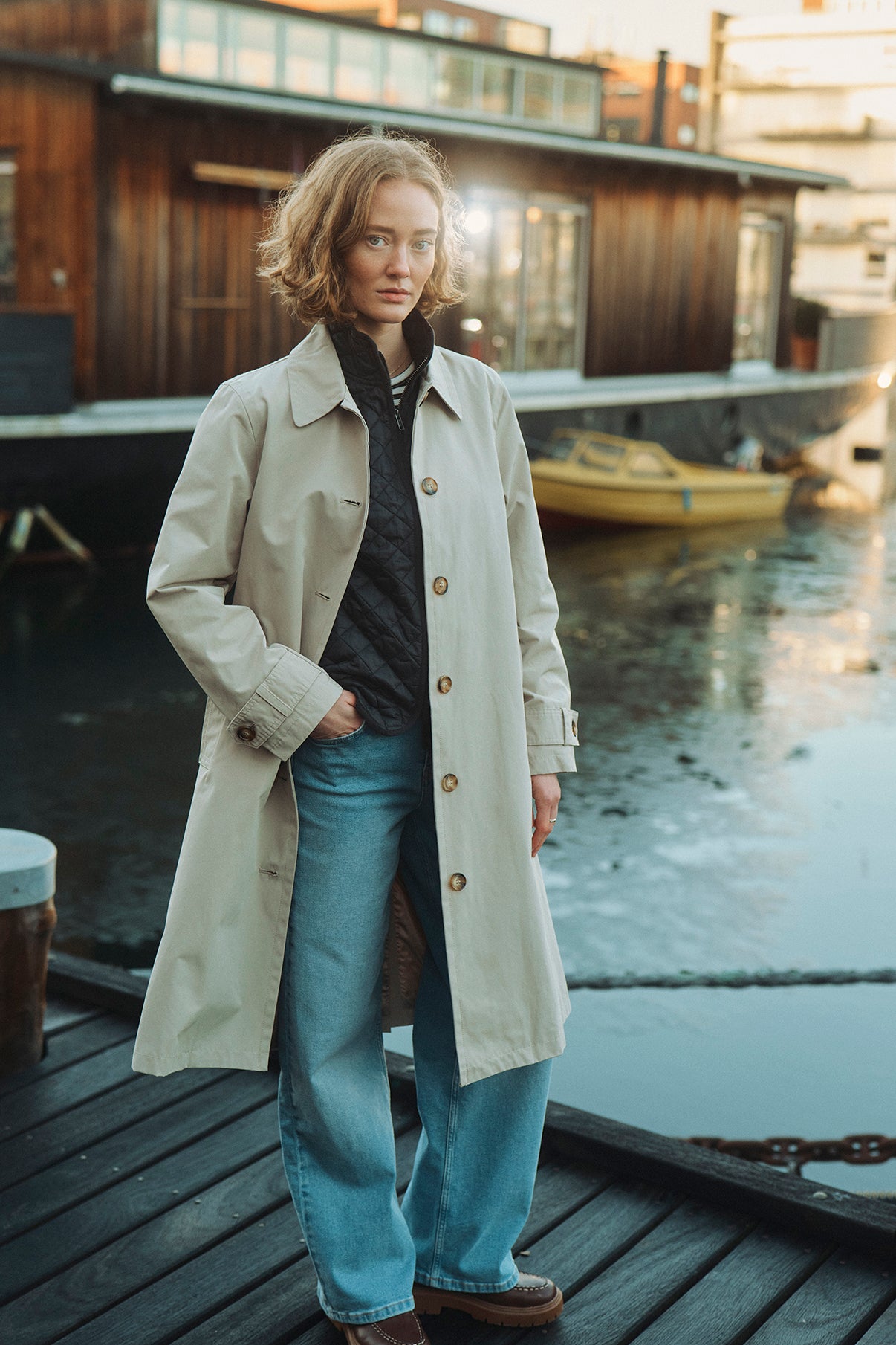 Lang beige trenchcoat med knapper og lommer, båret over sort quiltet vest og blå jeans.