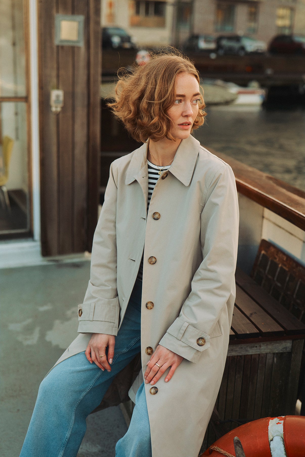 Oversize trenchcoat i lys beige med store knapper og lommer foran, stylet med blå jeans og stribet bluse.