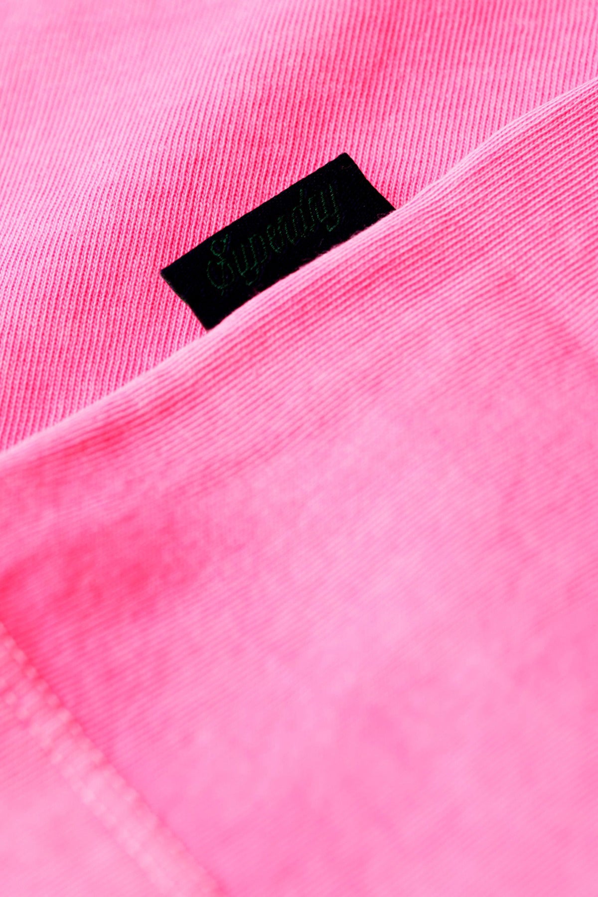 Pink neon T-shirt fabric close-up with small black rectangular logo patch and visible stitching.