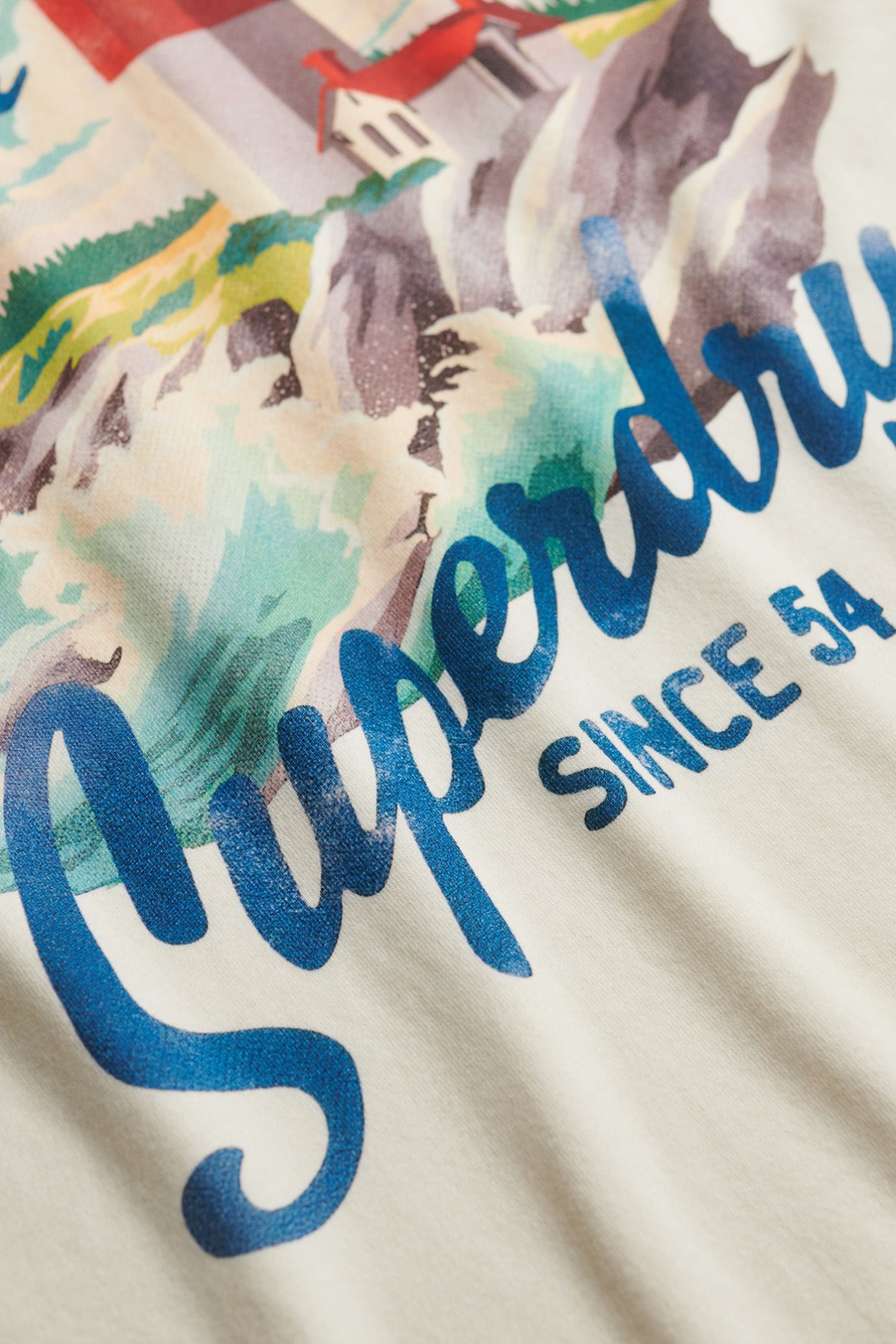 Detaljeret nærbillede af hvid kortærmet t-shirt med blå "Superdry Since 54"-tekst og farverigt grafisk print.