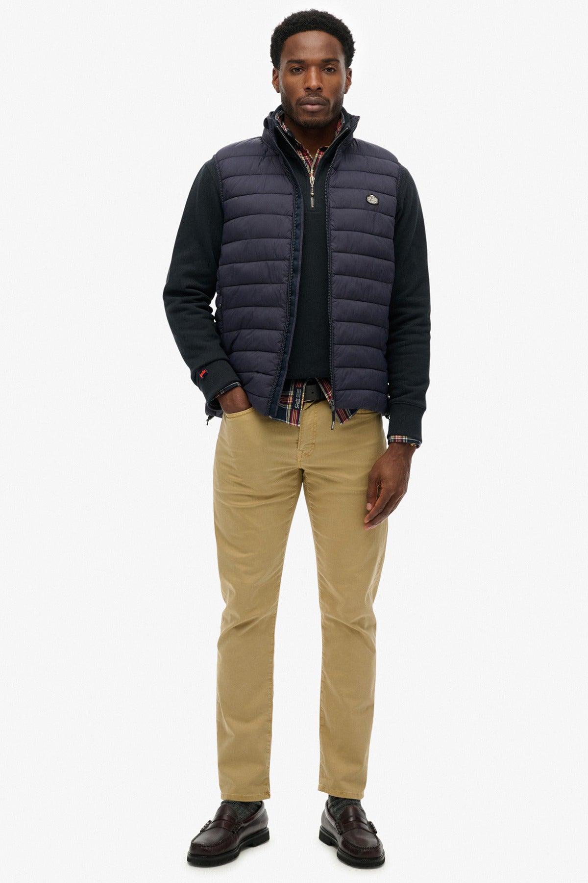 Mørkeblå, let polstret Superdry Fuji gilet med lynlås og slidser, båret over sort skjorte og beige bukser.