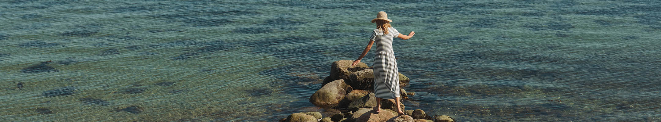Kvinde i stribet kjole med sommerhat på står på sten ved havet og ser ud over det klare vand