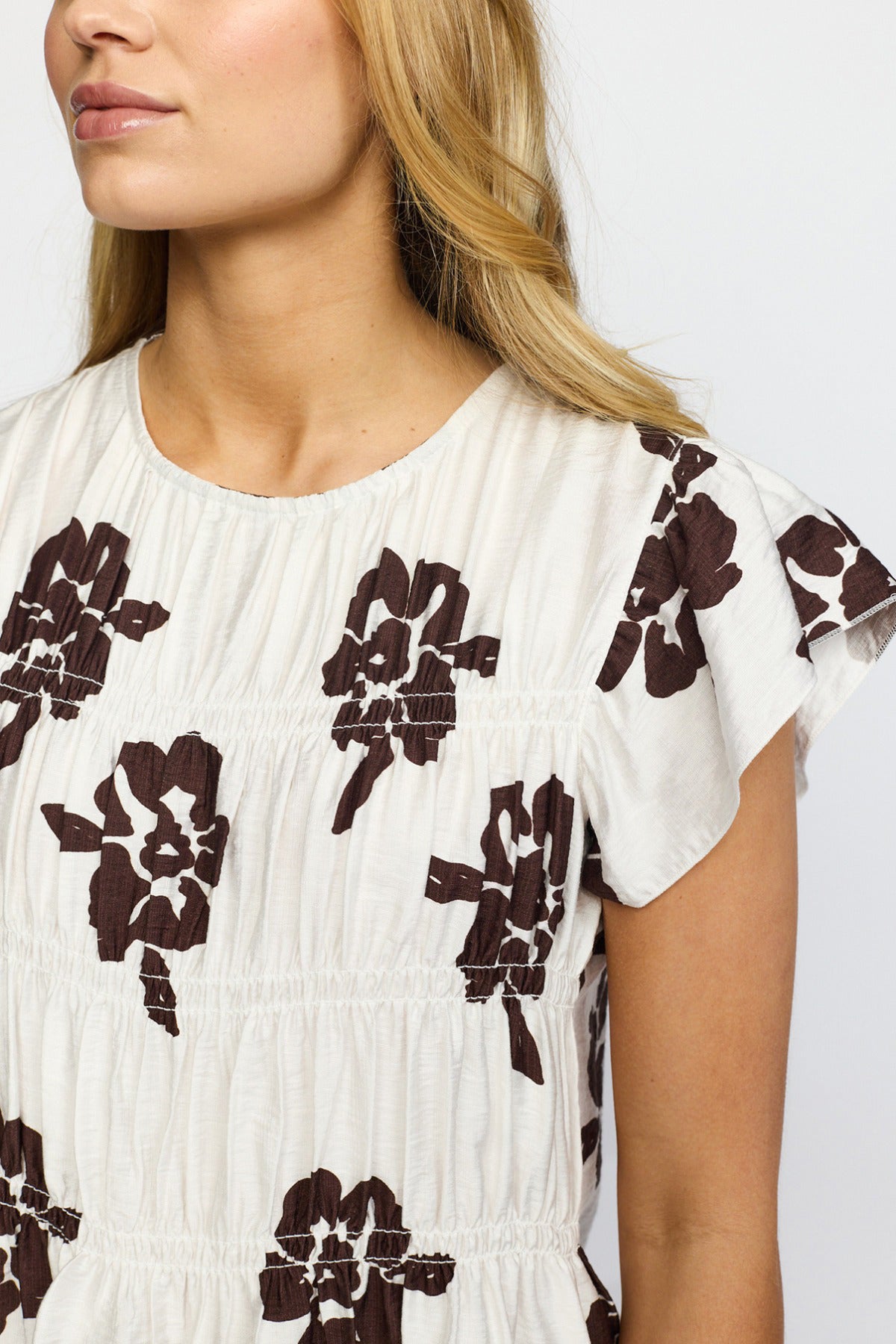Kortærmet hvid bluse med brune blomsterprint og rund hals med let rynkede detaljer.