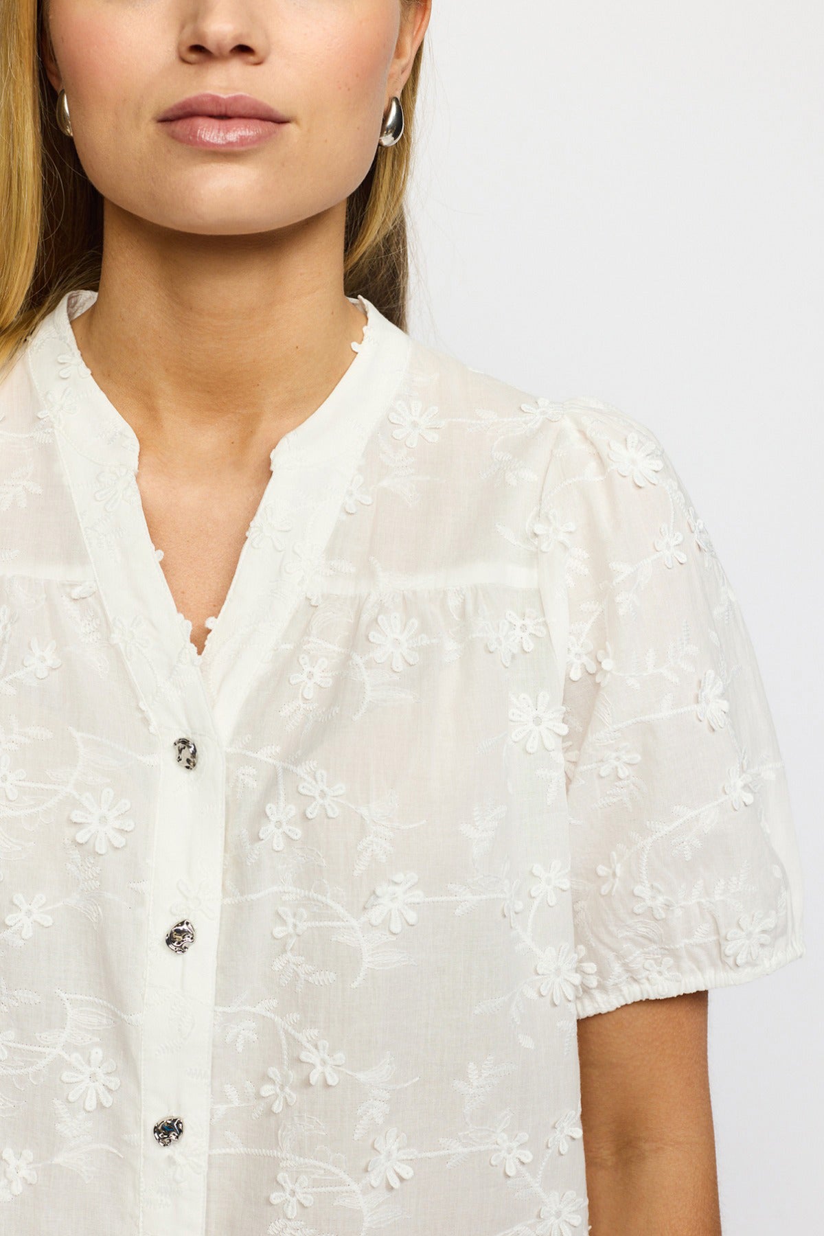 Hvid kortærmet bluse med gennemsigtigt blomsterrelief og små knapper foran.