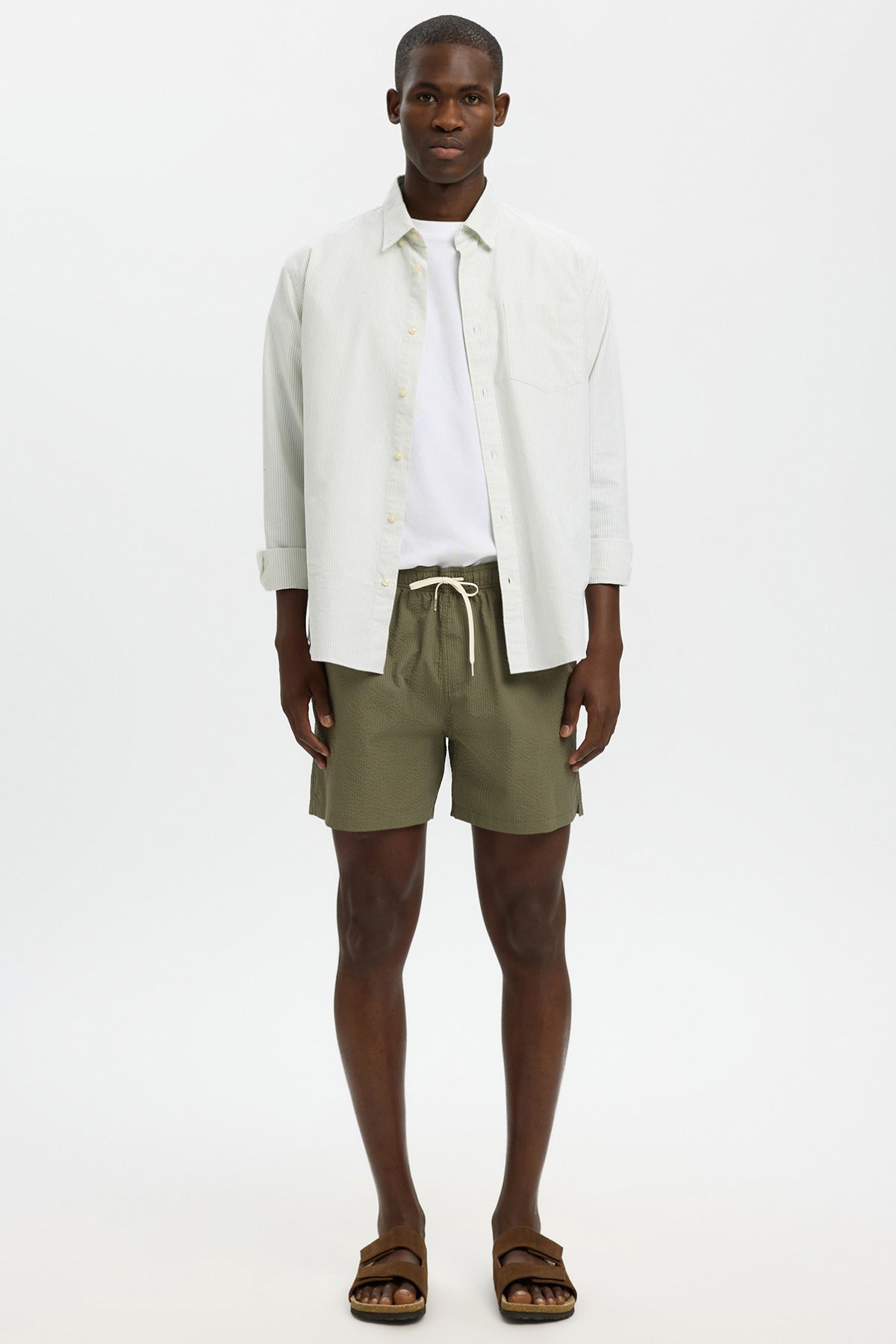 Herre badetøj i dyb lichen grøn med elastisk talje og snøre, kombineret med lys overshirt og sandfarvede sandaler.