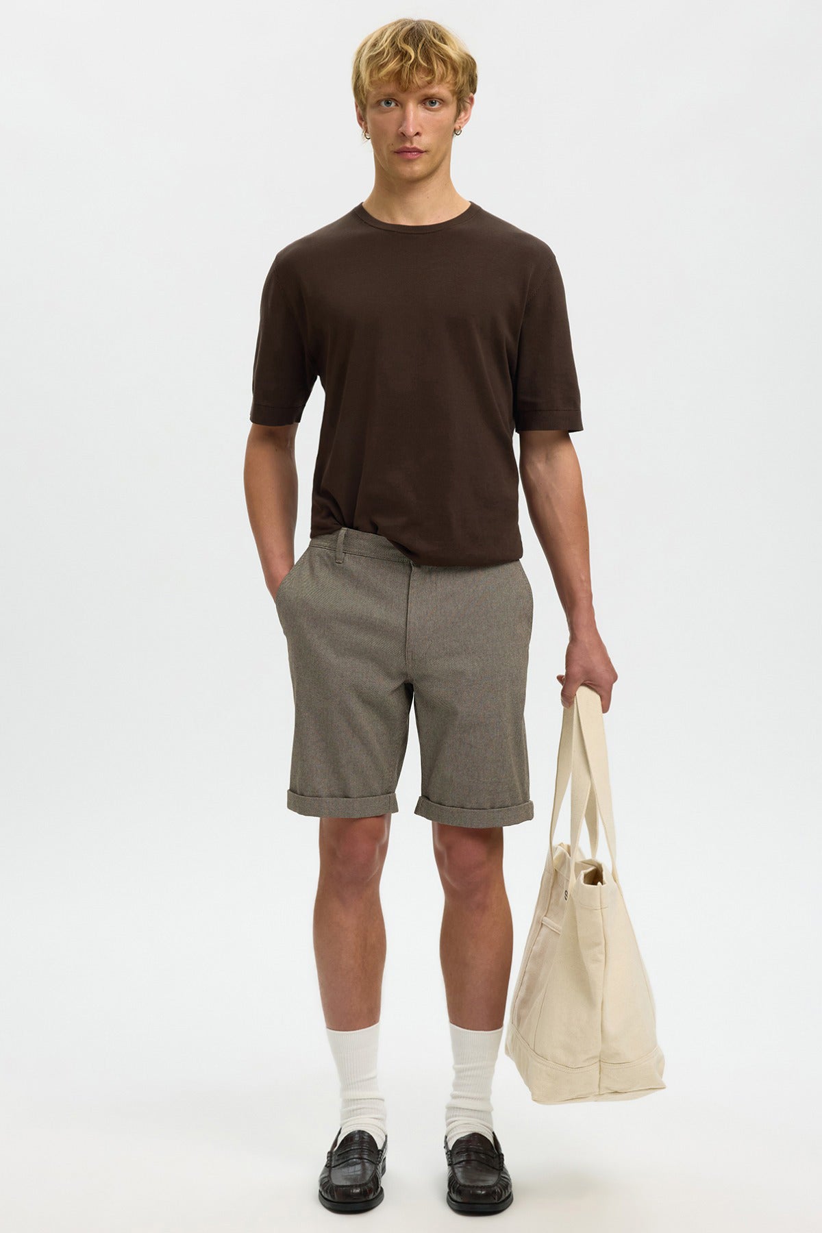 Mand iført grå strækbare shorts med lommer, mørkebrun t-shirt, hvide sokker og sorte sko, bærer stor beige indkøbstaske.