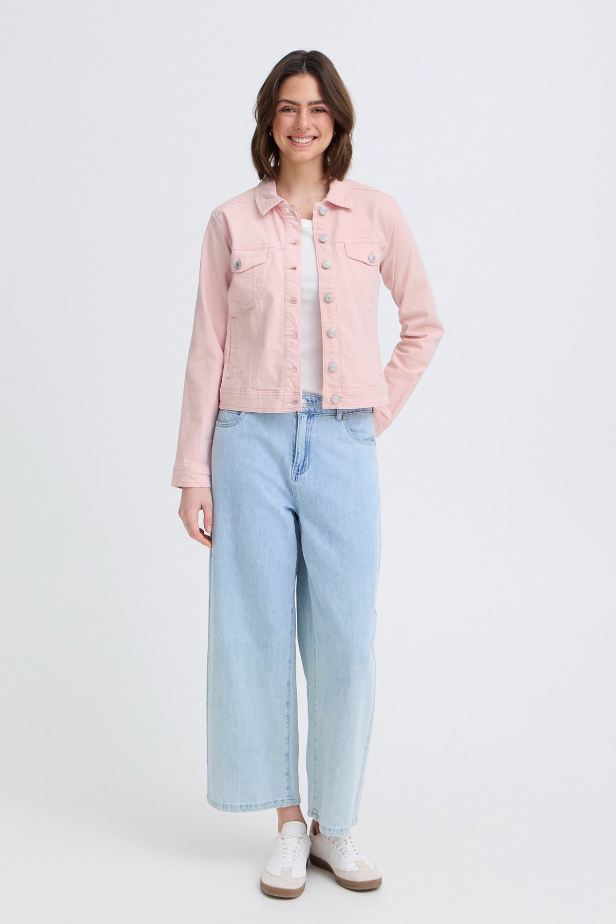 Lyserød elastisk denimjakke med knapper og brystlommer, model iført lyseblå jeans og hvide sko.