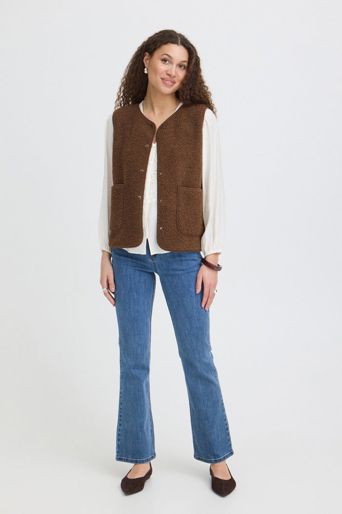 Brun teddyvest med knapper og rund hals på model med hvid bluse og blå jeans.