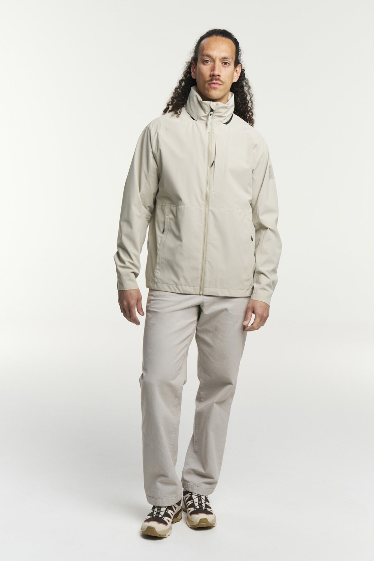 Beige funktionel skaljakke med høj krave og lang lynlås, kombineret med lyse bukser og sneakers.