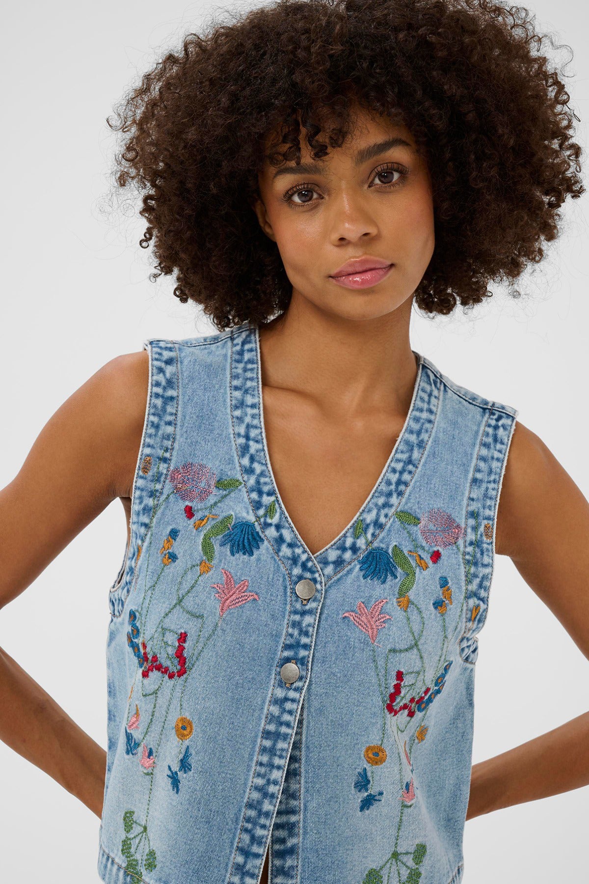 Lys blå denimvest med broderede blomsterdetaljer og V-udskæring uden ærmer.