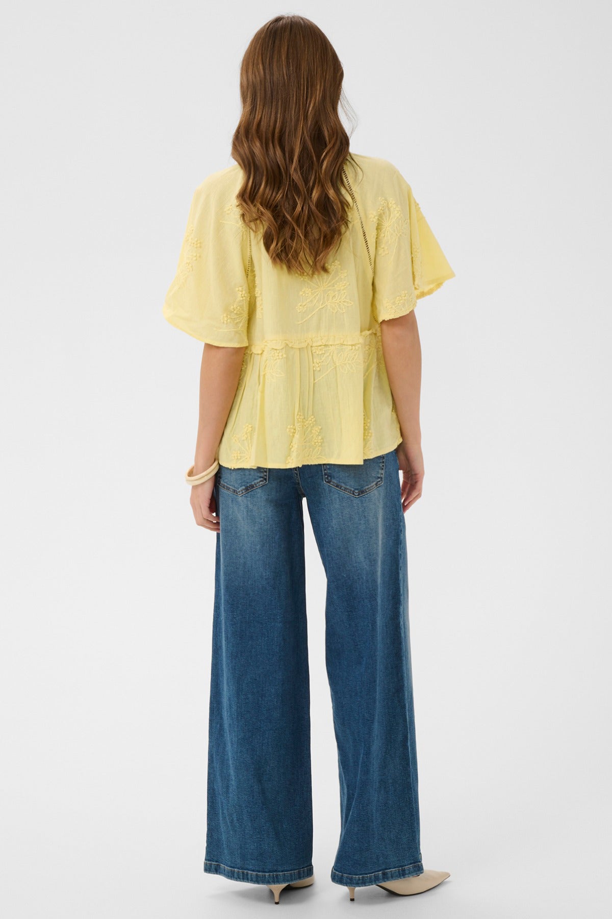Lemon-mørk gul kortærmet bluse med let, bølgende stof og broderi, set bagfra.