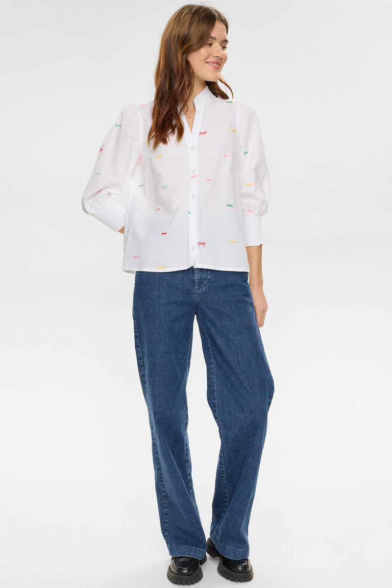 Hvid skjorte med farvede blomsterbroderier og ballonærmer på kvinde med blå jeans og sorte sko.
