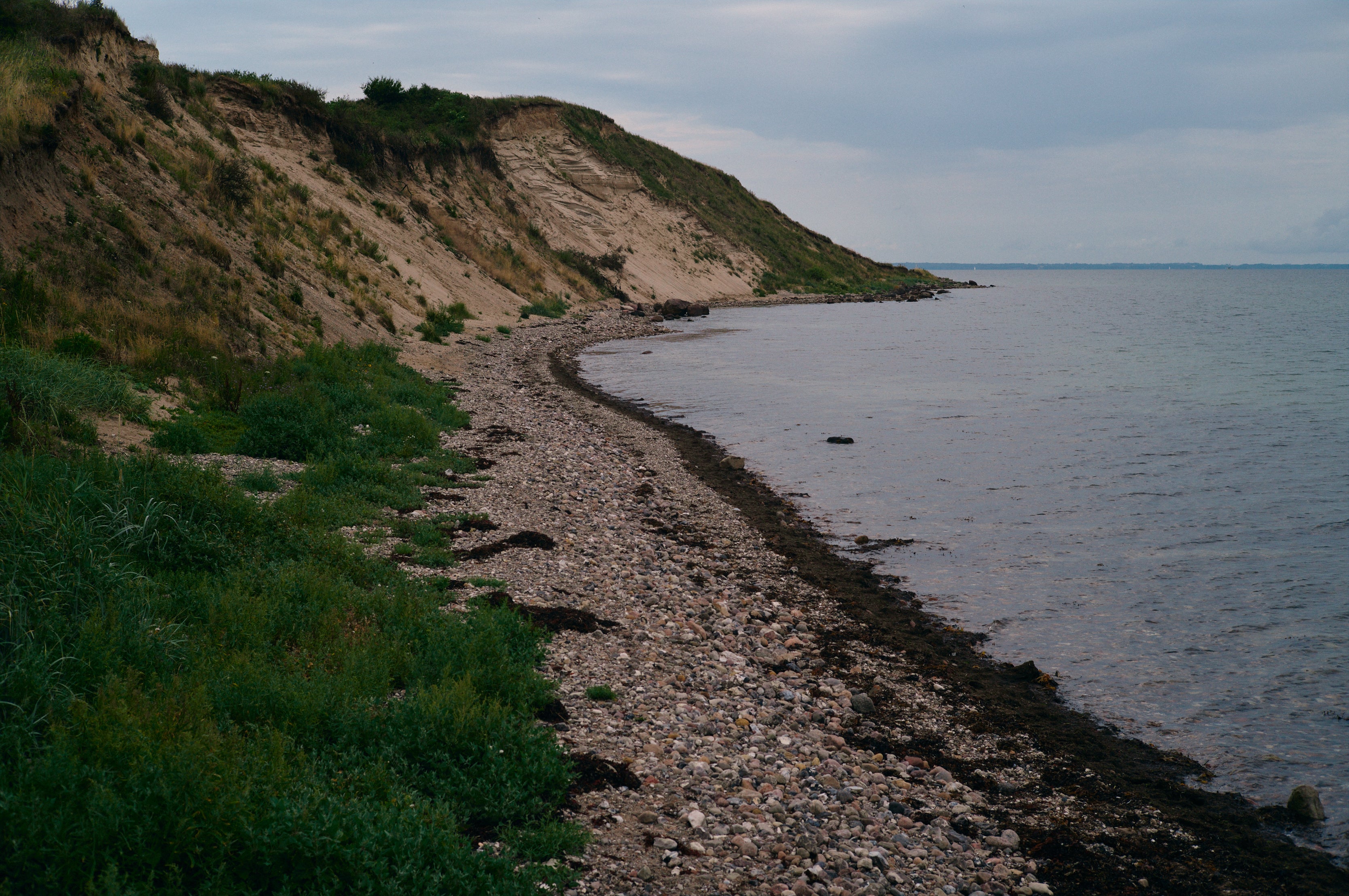 Kystlandskab med stenstrand, stejle klinter og roligt vand under en overskyet himmel.