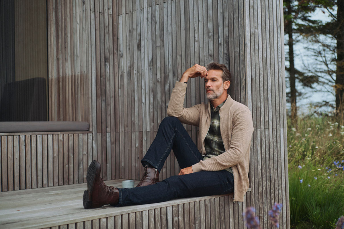 Mand i beige cardigan, ternet skjorte og mørke jeans sidder ved et træhus omgivet af natur.
