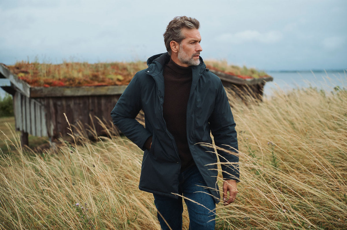 Mand i mørk jakke over en brun strik og jeans står i højt græs ved kysten med et træhus i baggrunden.