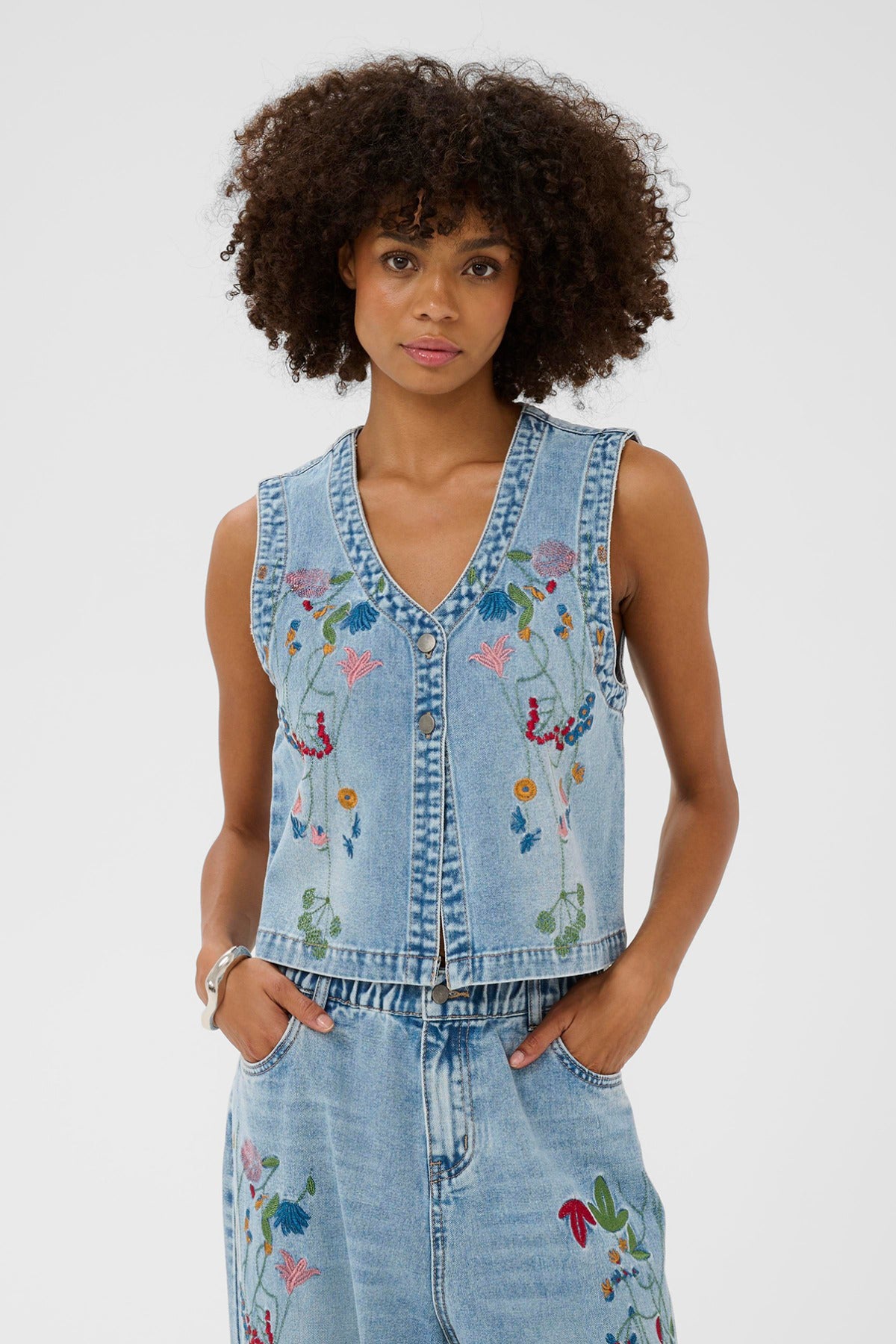 Lyseblå denimvest med farverig blomsterbrodering og knaplukning foran.