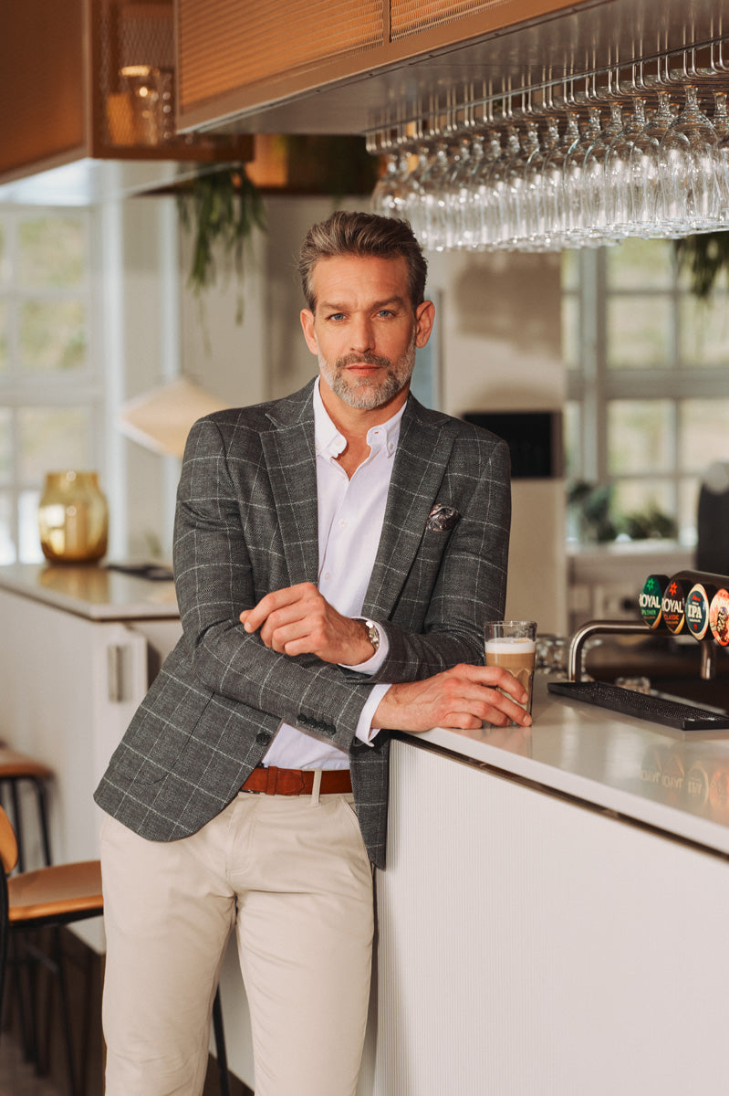 Mand i gråternet blazer, hvid skjorte og lyse chinos står ved en bar i et elegant smart casual look.