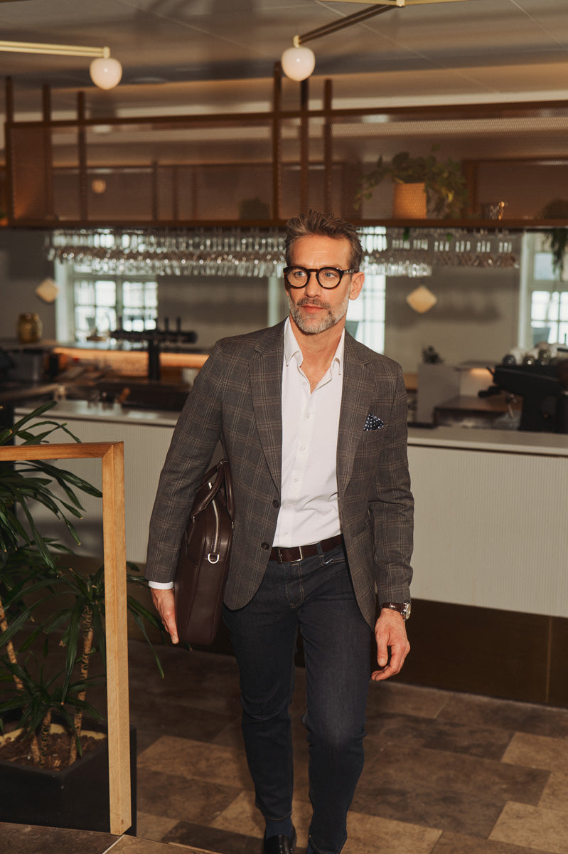 Mand i ternet blazer, hvid skjorte og mørke jeans med lædertaske i moderne business casual look.