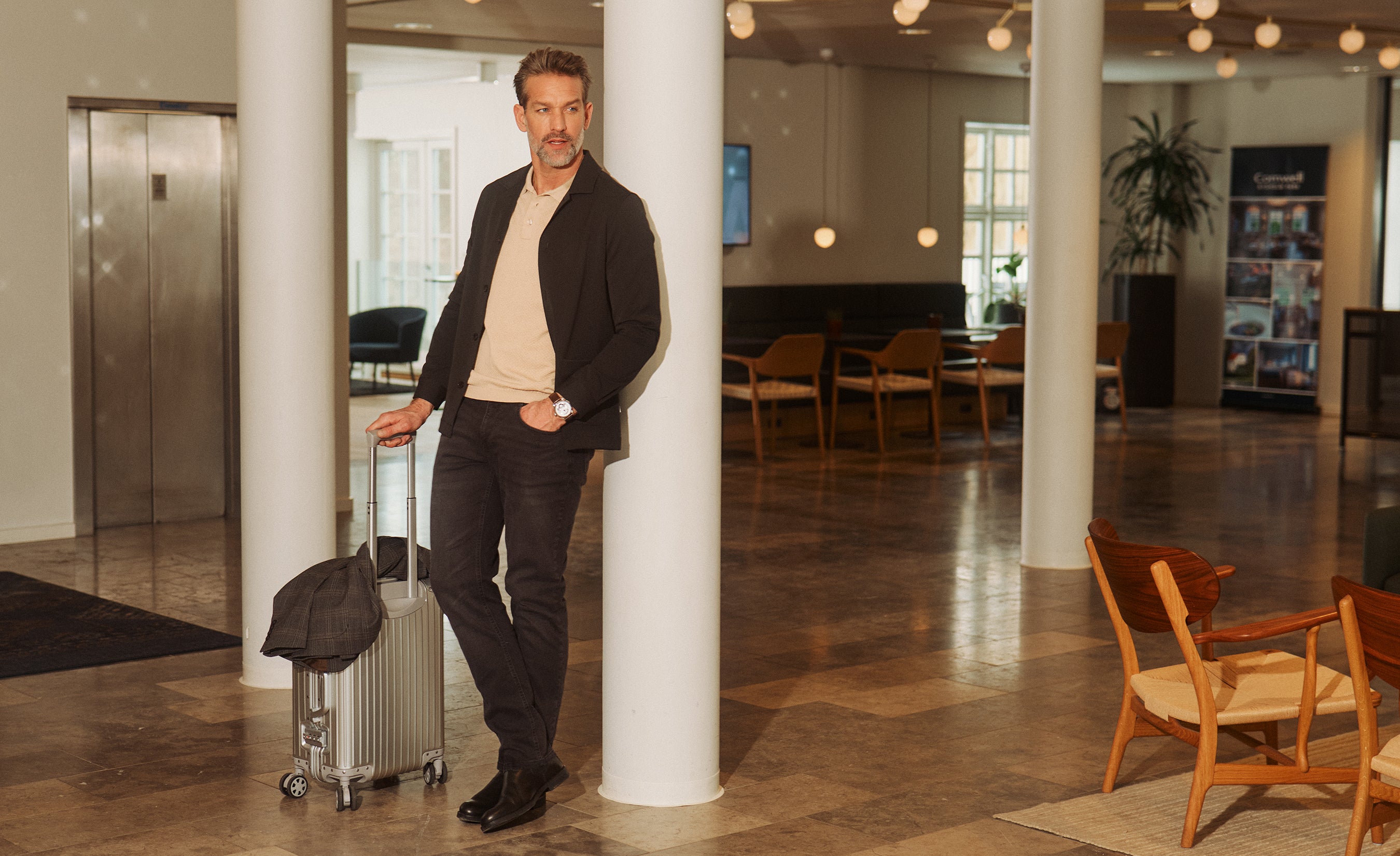 Mand i mørk overshirt, polo og jeans står med kuffert i moderne hotellobby og illustrerer et stilrent business casual look til rejse og arbejde.