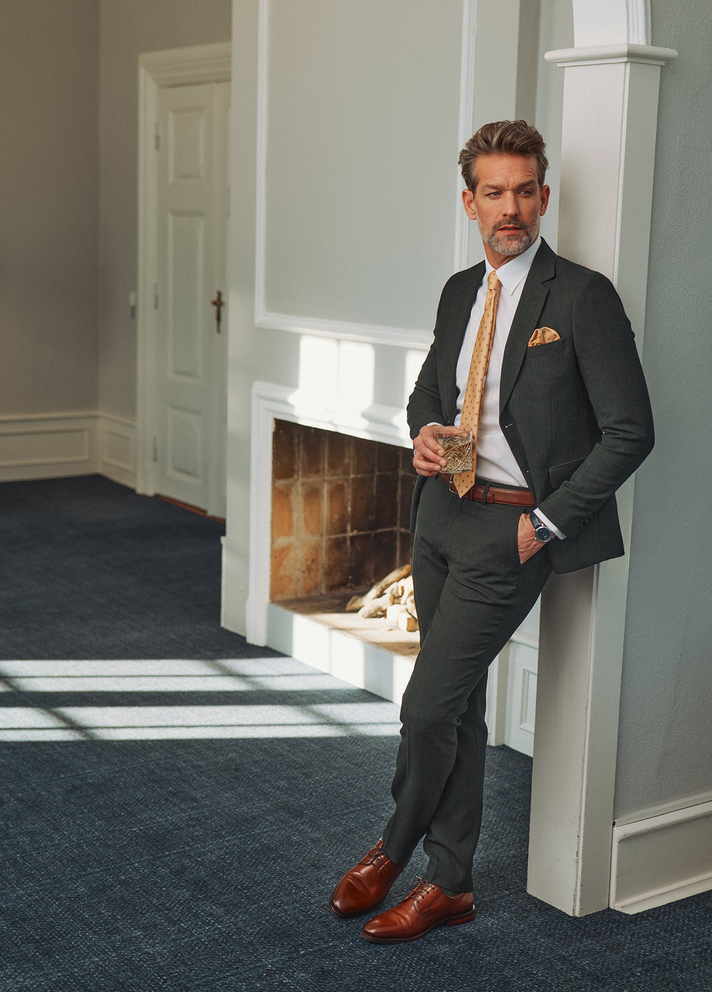 Mand i mørkegrønt jakkesæt med slips og brune lædersko står i elegante omgivelser med et klassisk og velklædt udtryk.