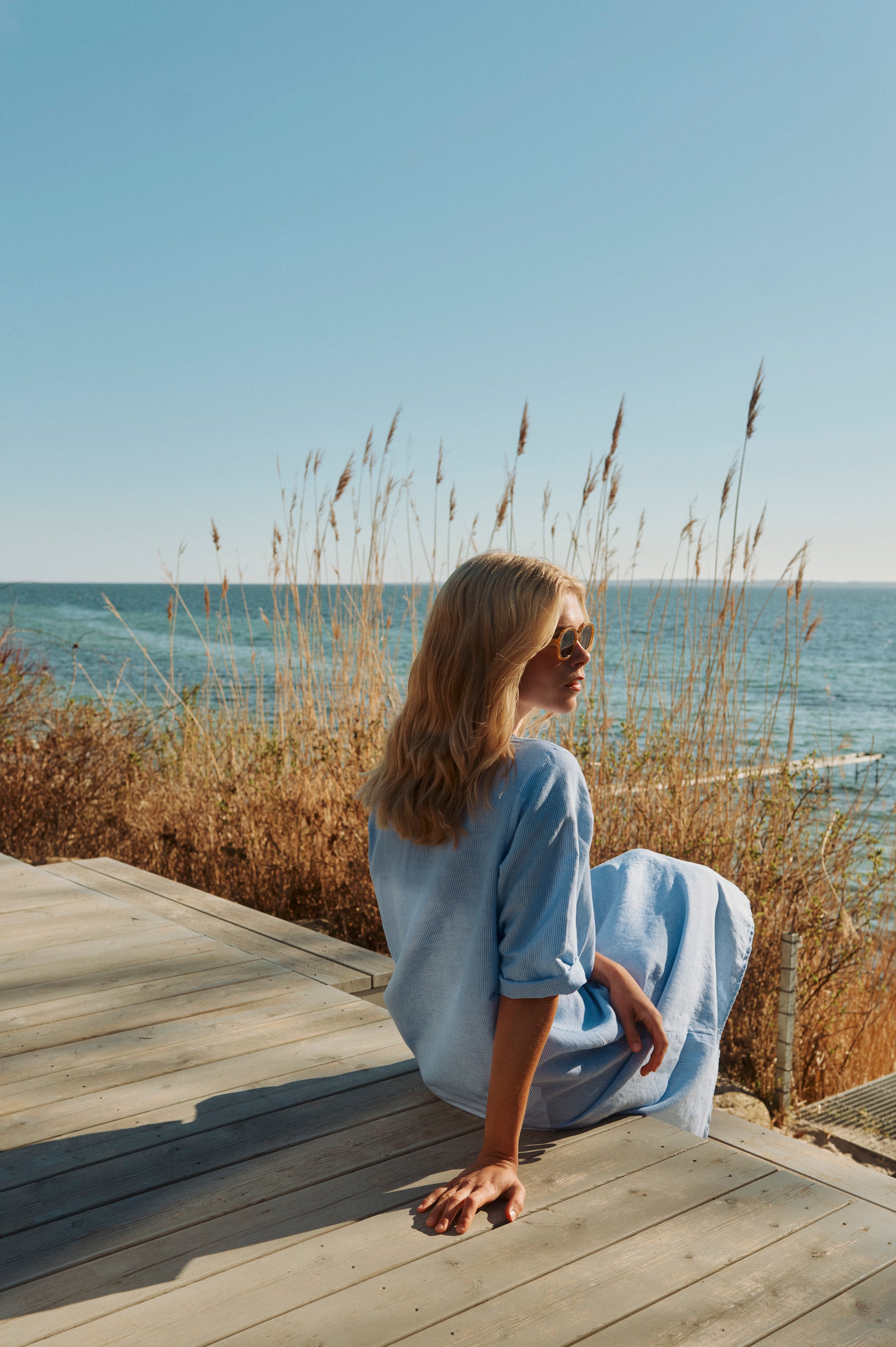 Kvinde i lyseblå sommerkjole sidder på en terrasse med udsigt over havet på en solrig dag
