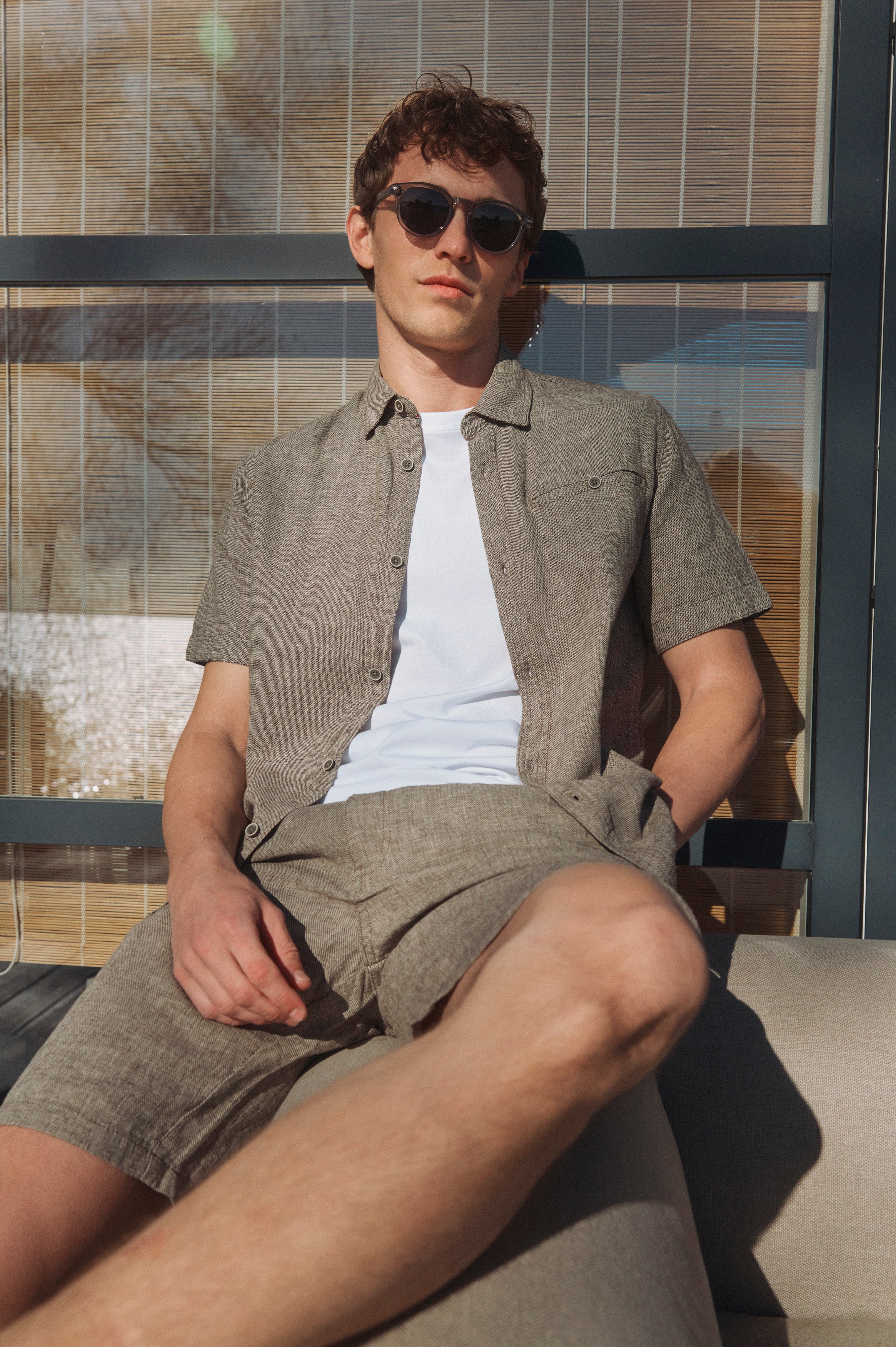 Mand i matchende hørskjorte og shorts sidder afslappet i solen med solbriller foran et sommerhus ved kysten.