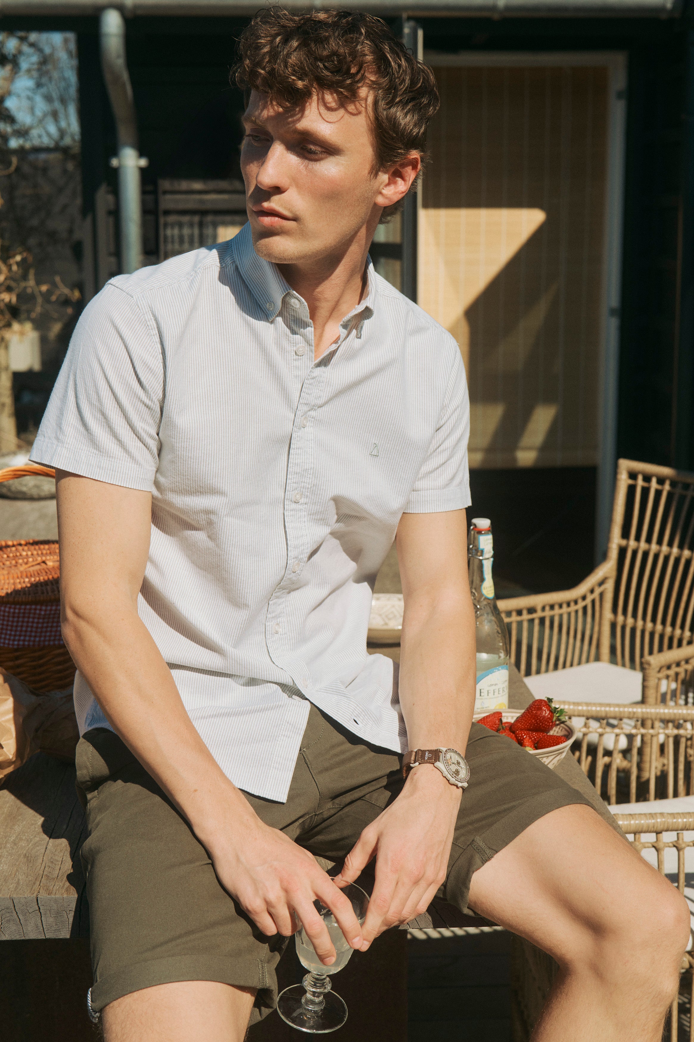 Afslappet sommerlook med kortærmet skjorte og shorts i lyse naturfarver på en solrig terrasse.