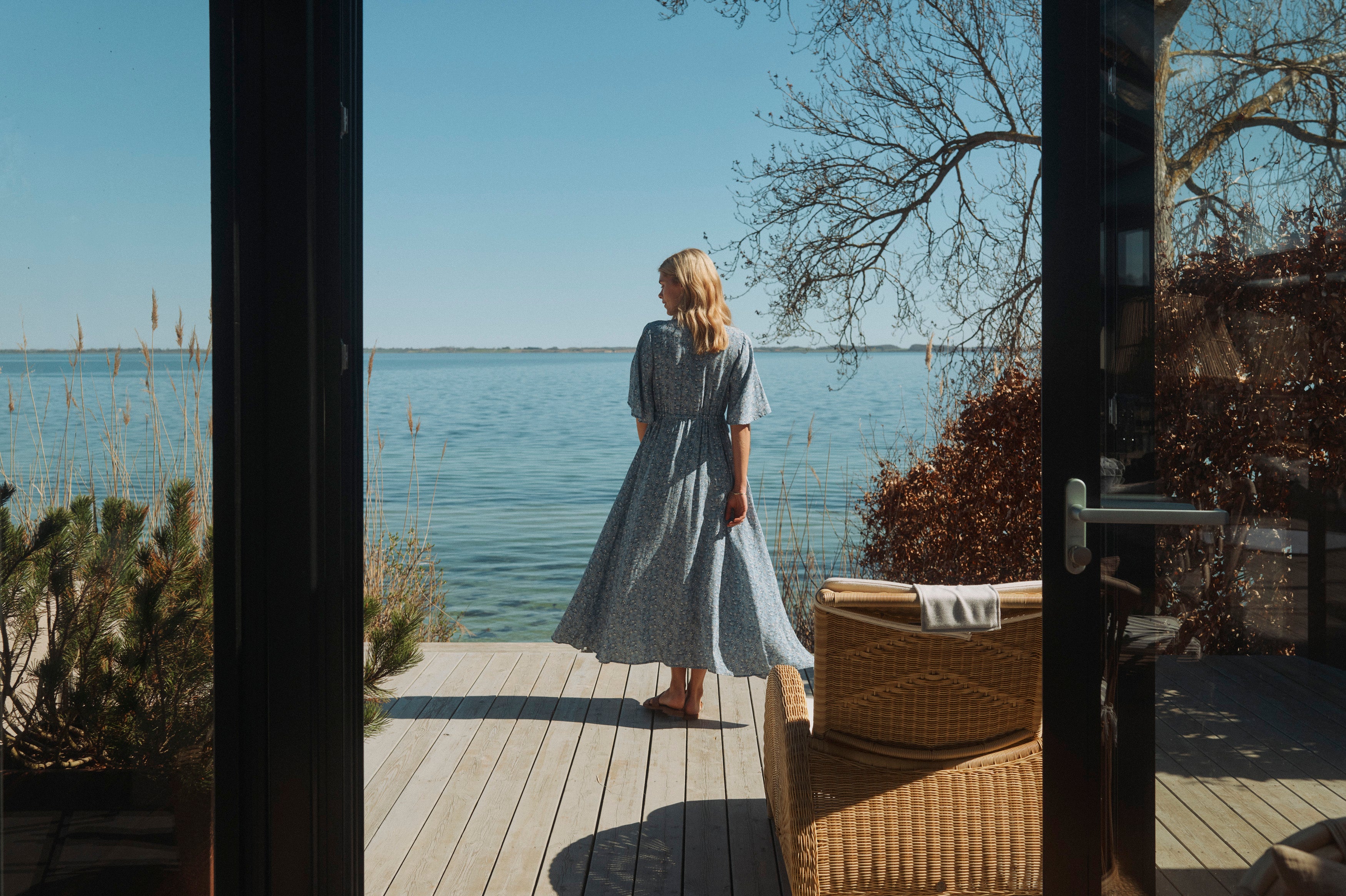 Kvinde i lyseblå blomsterkjole står på en terrasse med udsigt over havet på en solrig dag
