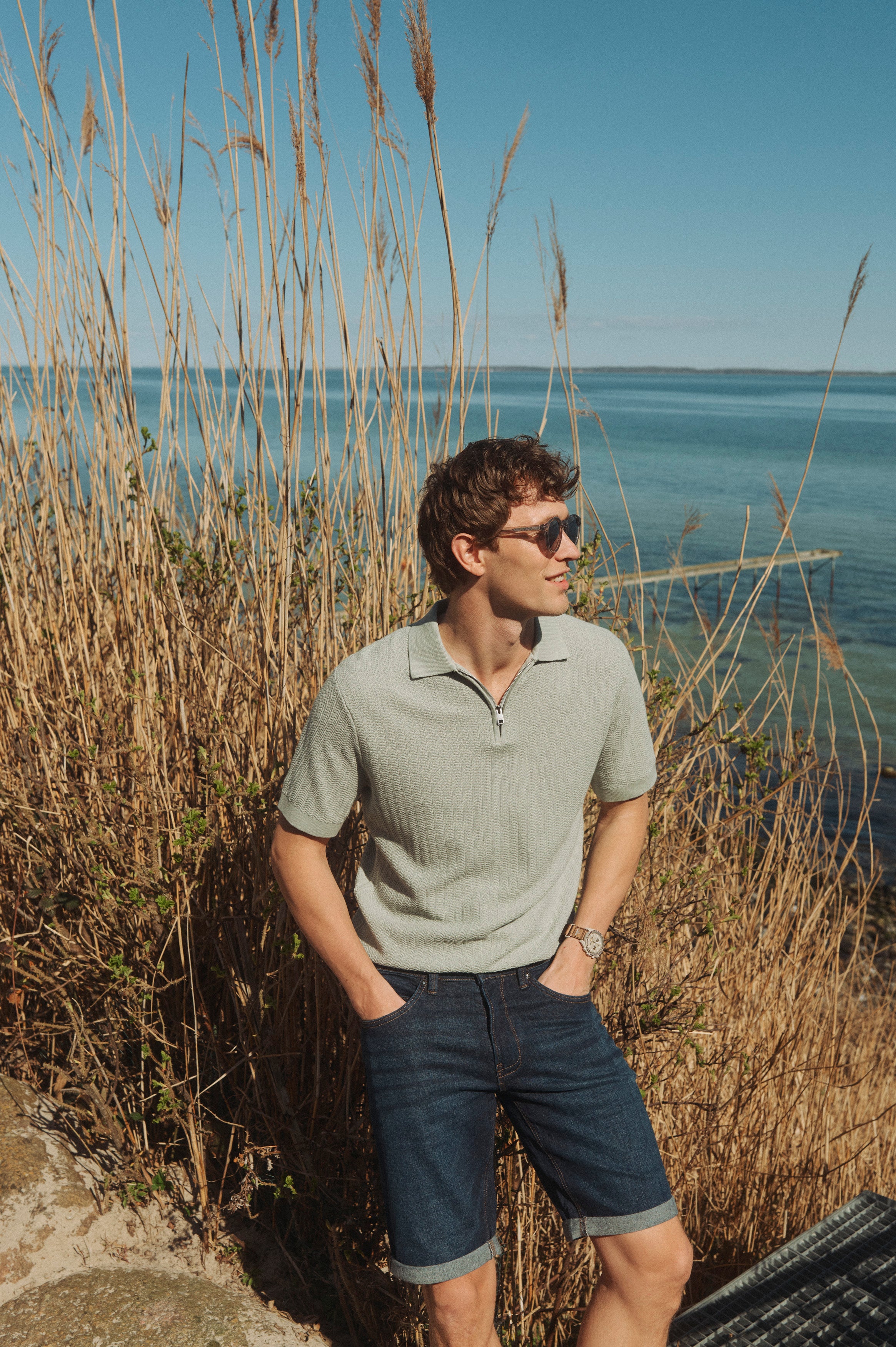 Mand i lysegrøn strikket polo og mørke denimshorts står ved siv og kystvand på en solrig sommerdag.