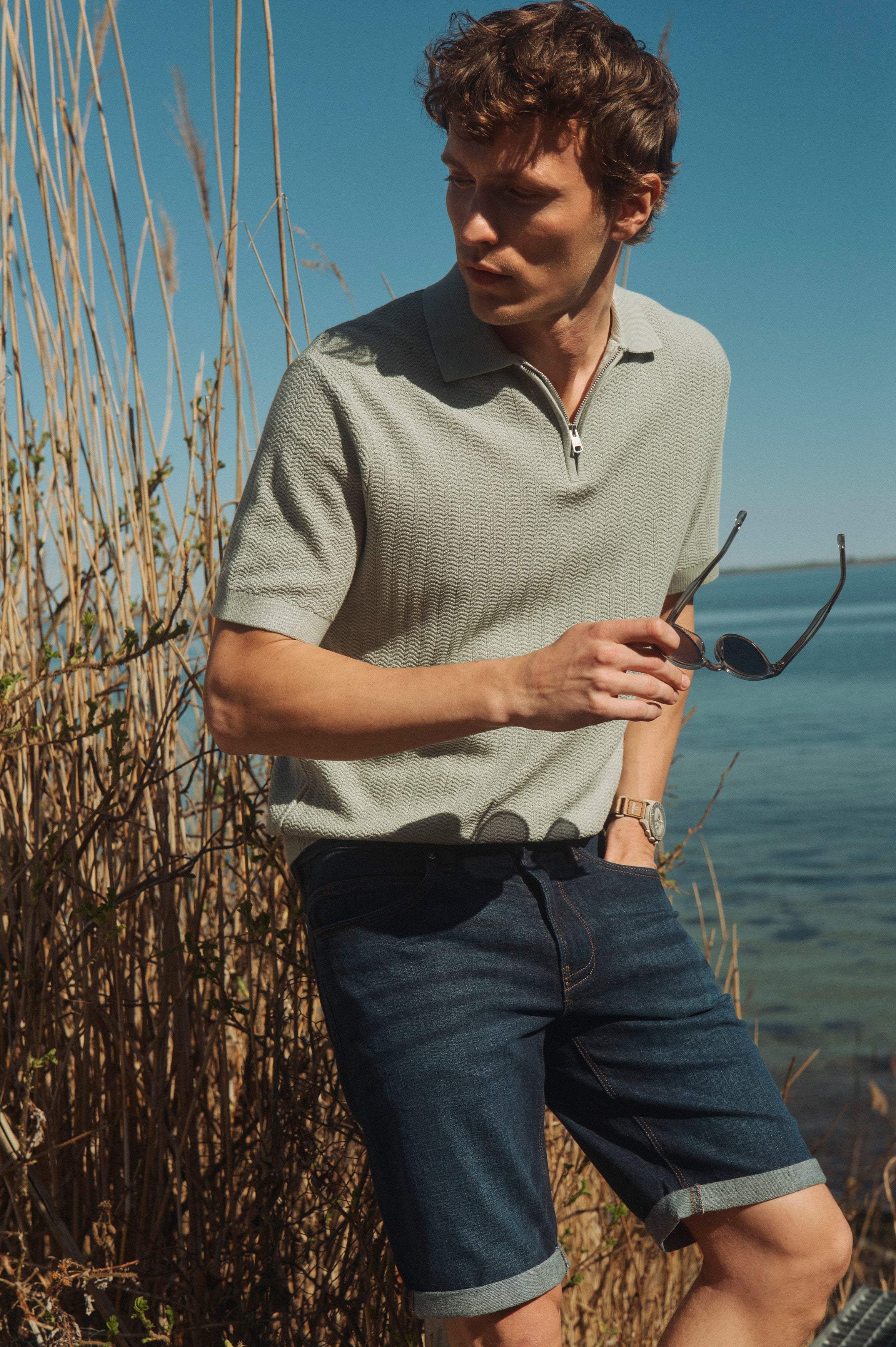 Mand i strikket polo og denimshorts står ved vandet med solbriller i hånden på en solrig sommerdag.