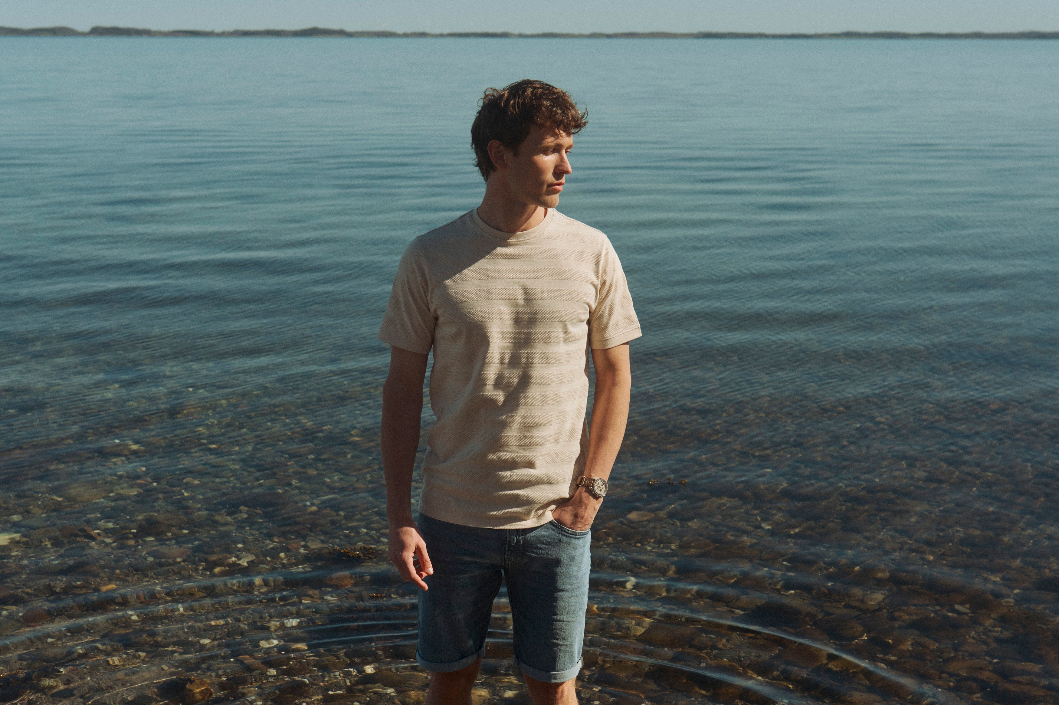 Mand i lys T-shirt og denimshorts står ved vandkanten på en solrig sommerdag med havet i baggrunden.