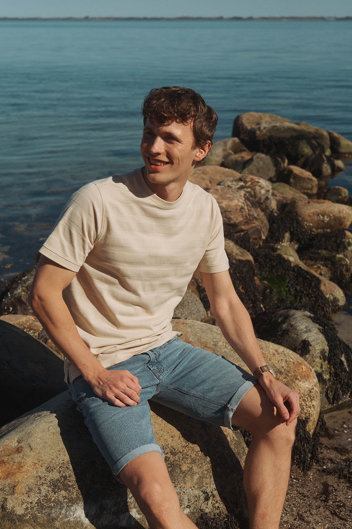 Herre i lyseblå denimshorts med opsmøgede kanter og beige t-shirt ved kysten.