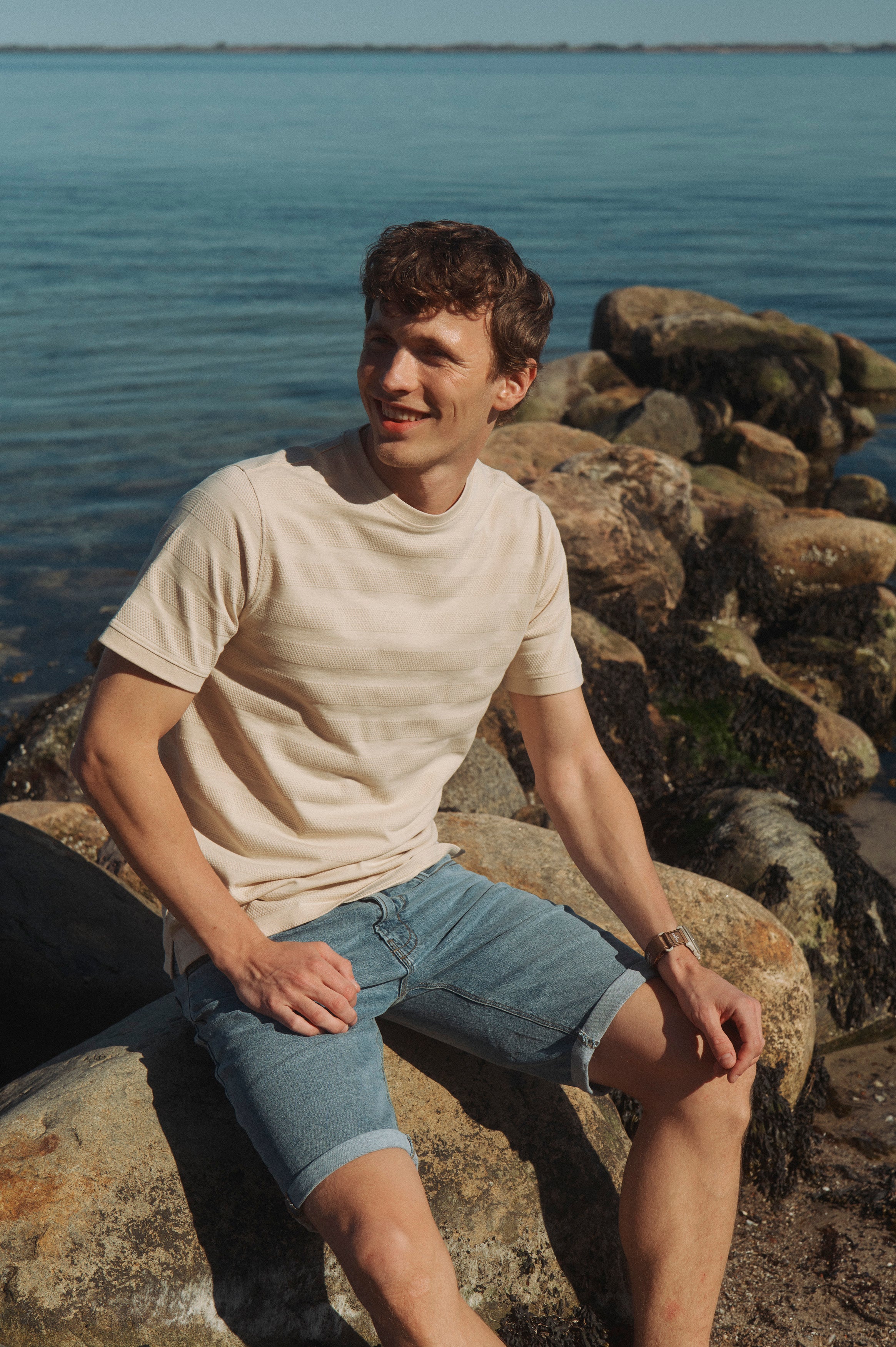 Mand i lys t-shirt og denimshorts sidder på klipper ved havet på en solrig sommerdag