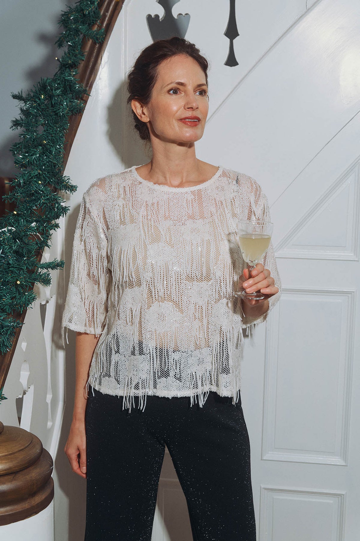 Kvinde i hvid festbluse med frynser og sorte glimmerbukser holder et glas ved julepyntet trappe – elegant tøj til julefrokost.