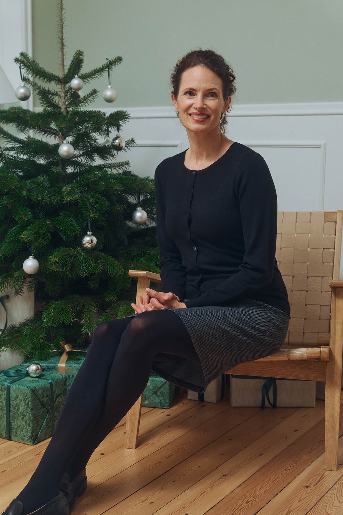 Kvinde i sort cardigan, grå nederdel og sorte strømpebukser sidder ved juletræet – klassisk og elegant tøj til juleaften.
