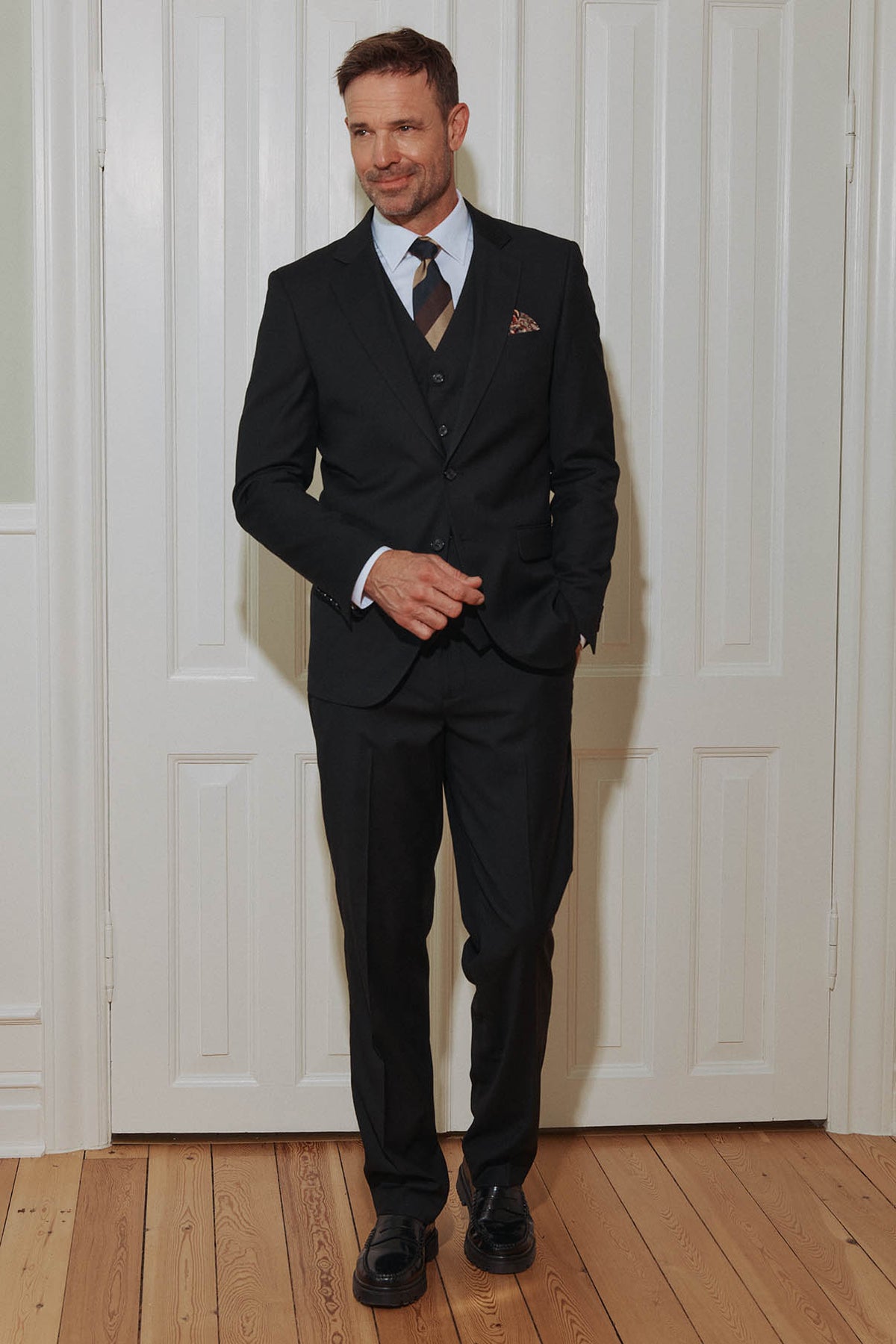 Mand i klassisk sort blazer med revers, hvid skjorte og slips, stående foran hvide døre.