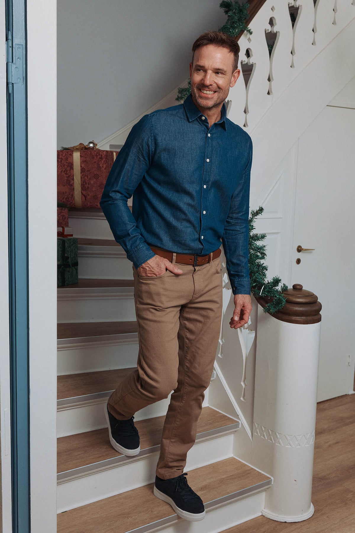 Mand i blå denimskjorte, beige bukser og sorte sneakers på julepyntet trappe – afslappet og stilfuldt herretøj til juleaften.
